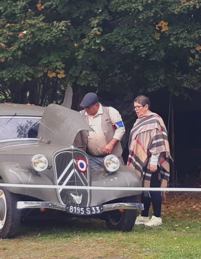 galerie,Trois personnes observent une voiture ancienne de la Seconde Guerre mondiale avec le capot ouvert. Le véhicule, une Citroën peinte en kaki avec l’inscription « FFI » (Forces françaises de l'intérieur), arbore un insigne tricolore sur la calandre. La scène se déroule dans un cadre arboré avec des tentes militaires à l’arrière, évoquant une reconstitution historique.