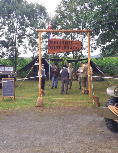 galerie, Entrée d’un campement militaire reconstitué avec une arche en bois portant l’inscription « Welcome to Fort Bragg ». Des personnes en tenue d’époque discutent à l’intérieur du camp, entouré de tentes militaires. Une jeep et un véhicule militaire sont visibles, ainsi qu’un tableau d’informations à l’entrée. L’environnement est rural, avec des arbres et un champ de maïs en arrière-plan.