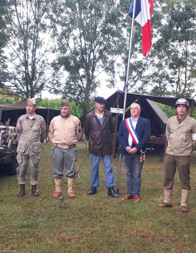 Un groupe de personnes en tenue d'époque se tient debout en extérieur devant une jeep militaire et des tentes de style militaire. La scène semble représenter une reconstitution historique, liée à la Seconde Guerre mondiale. Un homme porte une écharpe tricolore, suggérant un rôle officiel ou symbolique. Des drapeaux français sont visibles, renforçant le contexte historique et patriotique.