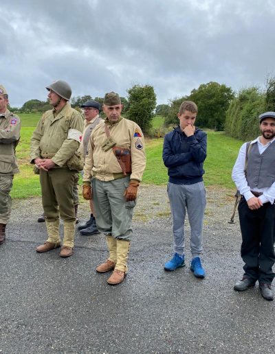 galerie, Des participants, certains en tenue militaire américaine de la Seconde Guerre mondiale et d’autres en civil, se tiennent en extérieur sur une route de campagne. Le ciel est nuageux. Parmi eux, un jeune homme porte une veste moderne contrastant avec les costumes historiques, tandis qu’un autre, en gilet et béret, évoque un résistant. À l’arrière-plan, quelques personnes âgées observent la scène.