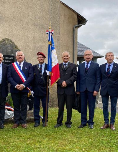 galerie, Dix hommes posent devant une stèle commémorative, certains en uniforme militaire ou en tenue officielle. L’un d’eux porte une écharpe tricolore, un autre tient un drapeau français d’anciens combattants. La scène a lieu devant un bâtiment, sur une pelouse avec vue sur la campagne. Il s'agit d'une cérémonie d'hommage aux anciens combattants ou victimes de guerre à Lansac.