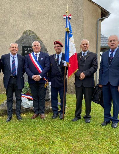 galerie, Sept hommes en costume ou tenue officielle posent devant une stèle commémorative à Lansac. L’un d’eux porte une écharpe tricolore de maire, un autre un béret rouge et tient un drapeau français d’anciens combattants. Une gerbe a été déposée au pied de la stèle. d’hommage aux morts pour la France.