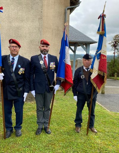 galerie, Trois porte-drapeaux en tenue d’anciens combattants posent devant une stèle commémorative. Deux portent un béret rouge et de nombreuses décorations, le troisième un béret bleu marine. Chacun tient un drapeau tricolore orné de franges dorées. L’arrière-plan montre un paysage vallonné sous un ciel nuageux,