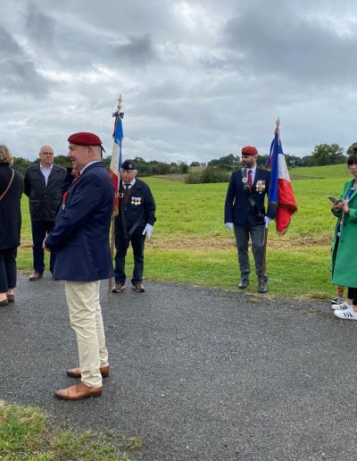 galerie, Des participants à la cérémonie mémorielle se tiennent en extérieur sur une route bordée de champs. Plusieurs hommes portent des décorations et des bérets rouges ou bleus, tenant des drapeaux tricolores. D'autres personnes, en tenue civile, discutent ou observent la scène, tandis qu'une femme en manteau vert consulte son téléphone. Le ciel est couvert, créant une atmosphère solennelle.